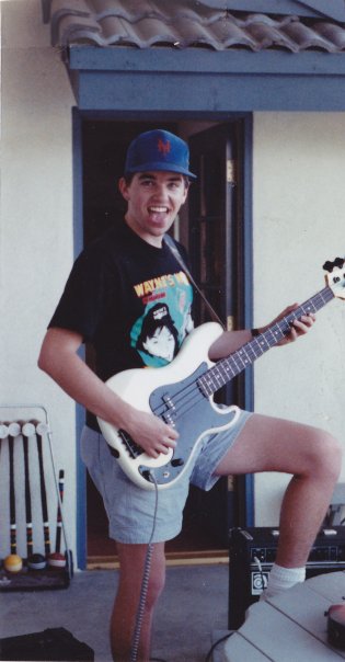 Brian playing a white bass with a Wayne's World shirt and a Mets hat on wearing shorts