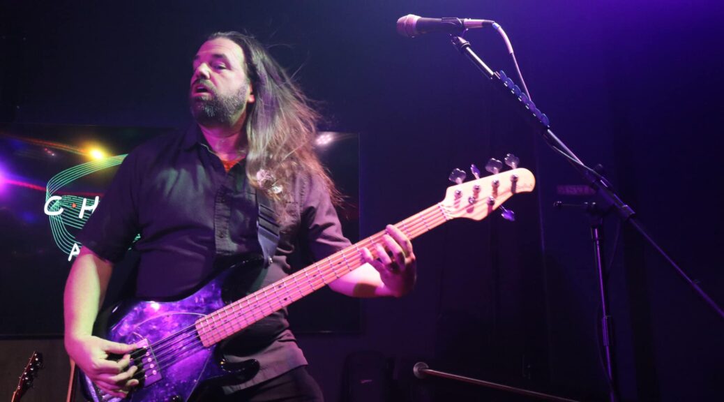 Handsome long-haired male bassist with a beard playing live on stage with his microphone visible