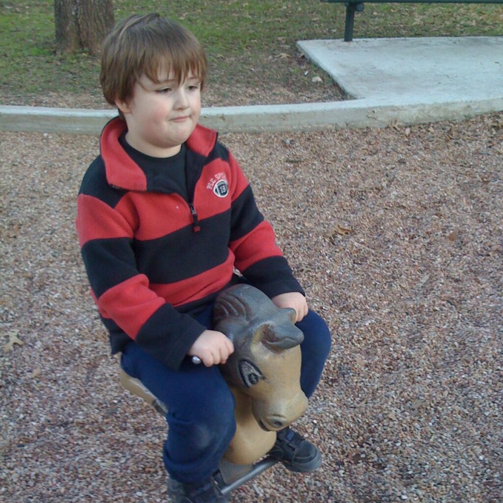 A bored-looking boy in a red and black striped pullover jacket and jeans rides a beaten-up playground horse that is too small for him.